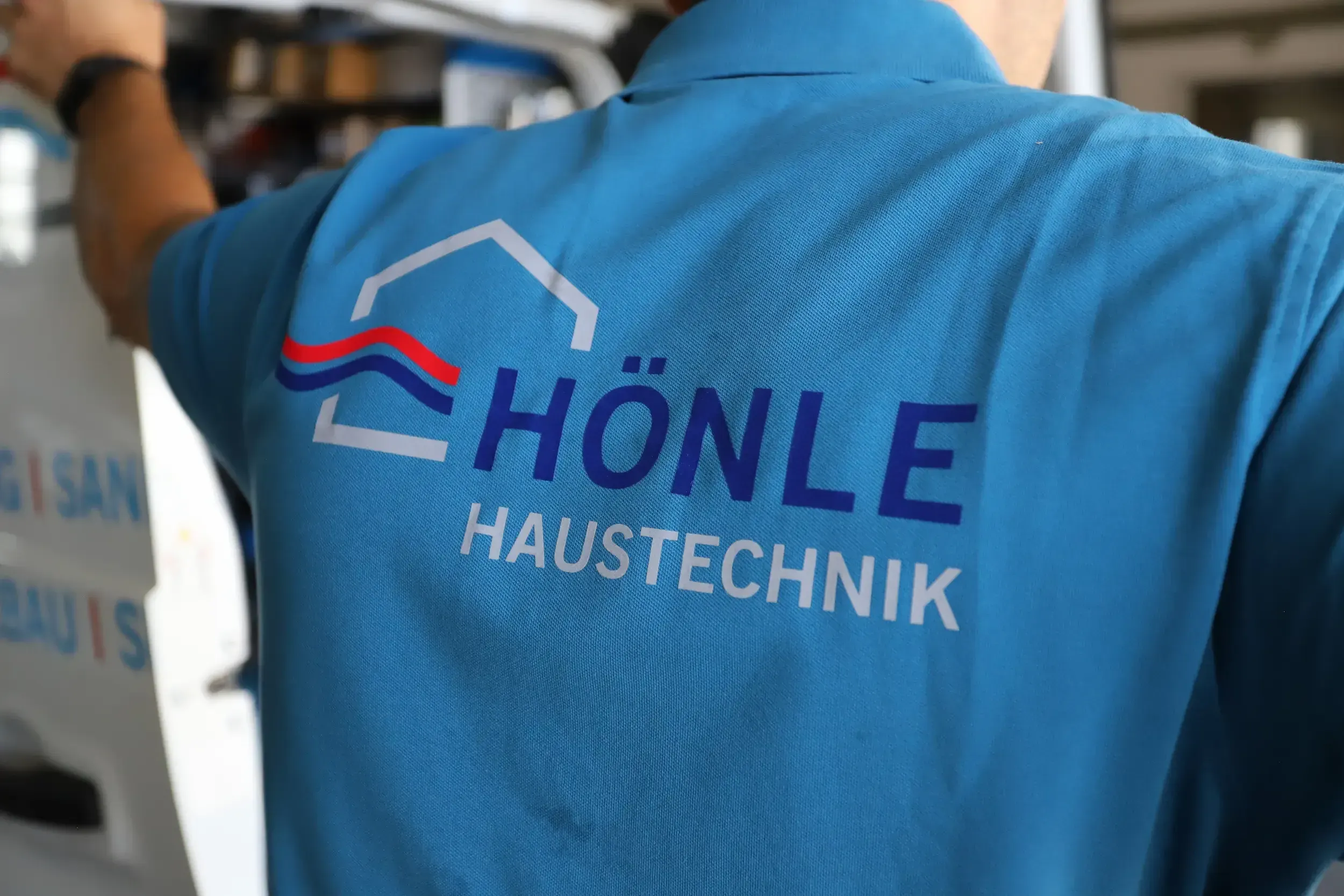 Mitarbeiter von Hönle Haustechnik von hinten im blauen Poloshirt mit Firmenlogo auf dem Rücken, am geöffneten Firmenwagen